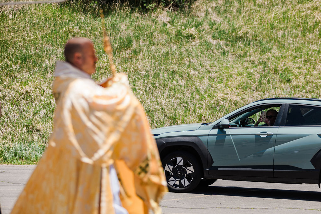 Blurred priest in gold robes stands by a green SUV as the driver takes a photo on a sunny roadside with grassy hill behind.