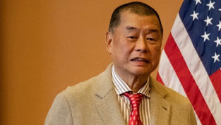 Man in a beige suit and red tie stands in front of a U.S. flag, with a neutral expression in an indoor setting.
