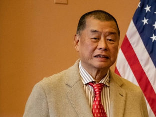 Man in a beige suit and red tie stands in front of a U.S. flag, with a neutral expression in an indoor setting.