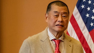 Man in a beige suit and red tie stands in front of a U.S. flag, with a neutral expression in an indoor setting.