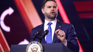 Man in a suit speaks at podium with "Vice President of the United States" seal. Red and orange lights in the background. Determined expression.