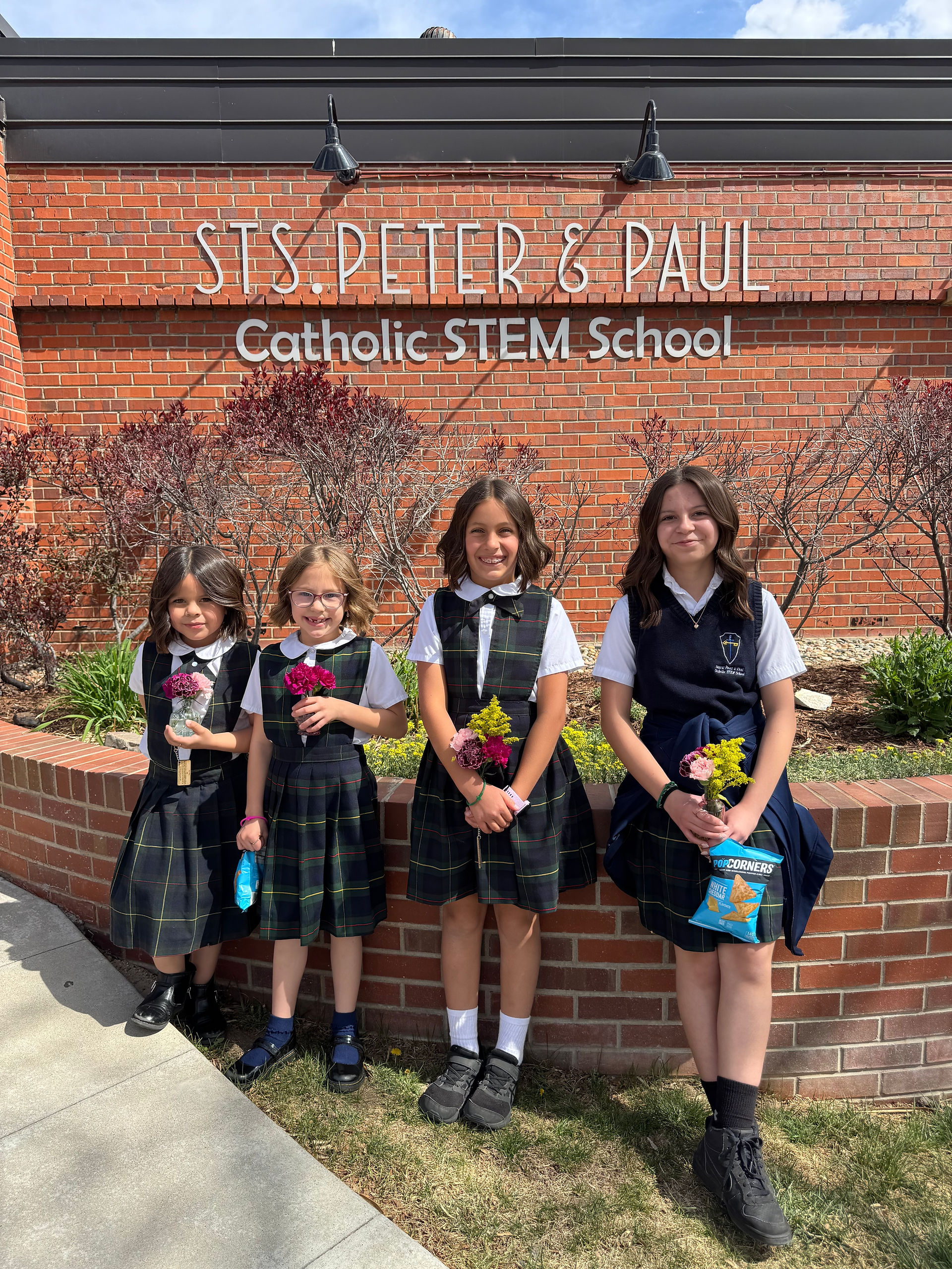 Four schoolgirls in uniform stand outside a brick building labeled ‘Sts. Peter & Paul Catholic STEM School,’ holding small bouquets of flowers.