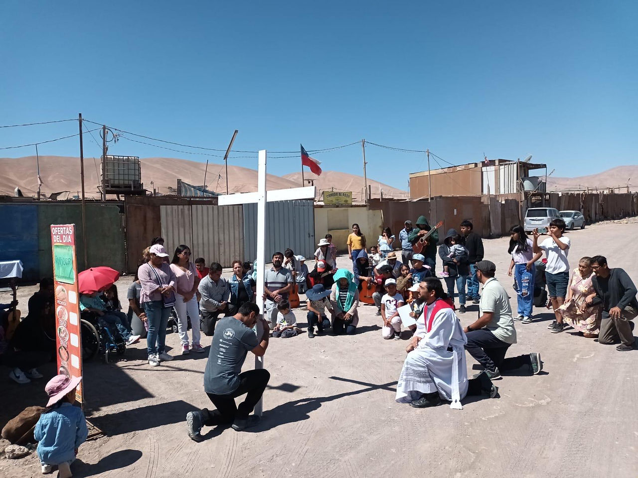 A group of people kneels in a desert setting under a clear blue sky, with a man in religious attire. A large cross is present. Mood is solemn.