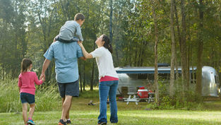 Family walking in a forest, with a boy on a man's shoulders and a girl holding his hand. A trailer and picnic table are in the background.