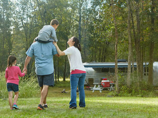 Family walking in a forest, with a boy on a man's shoulders and a girl holding his hand. A trailer and picnic table are in the background.