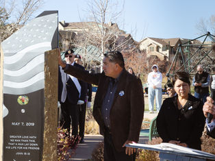 Man touches a stone memorial with an American flag design and the date May 7, 2019. People stand solemnly in a park setting.