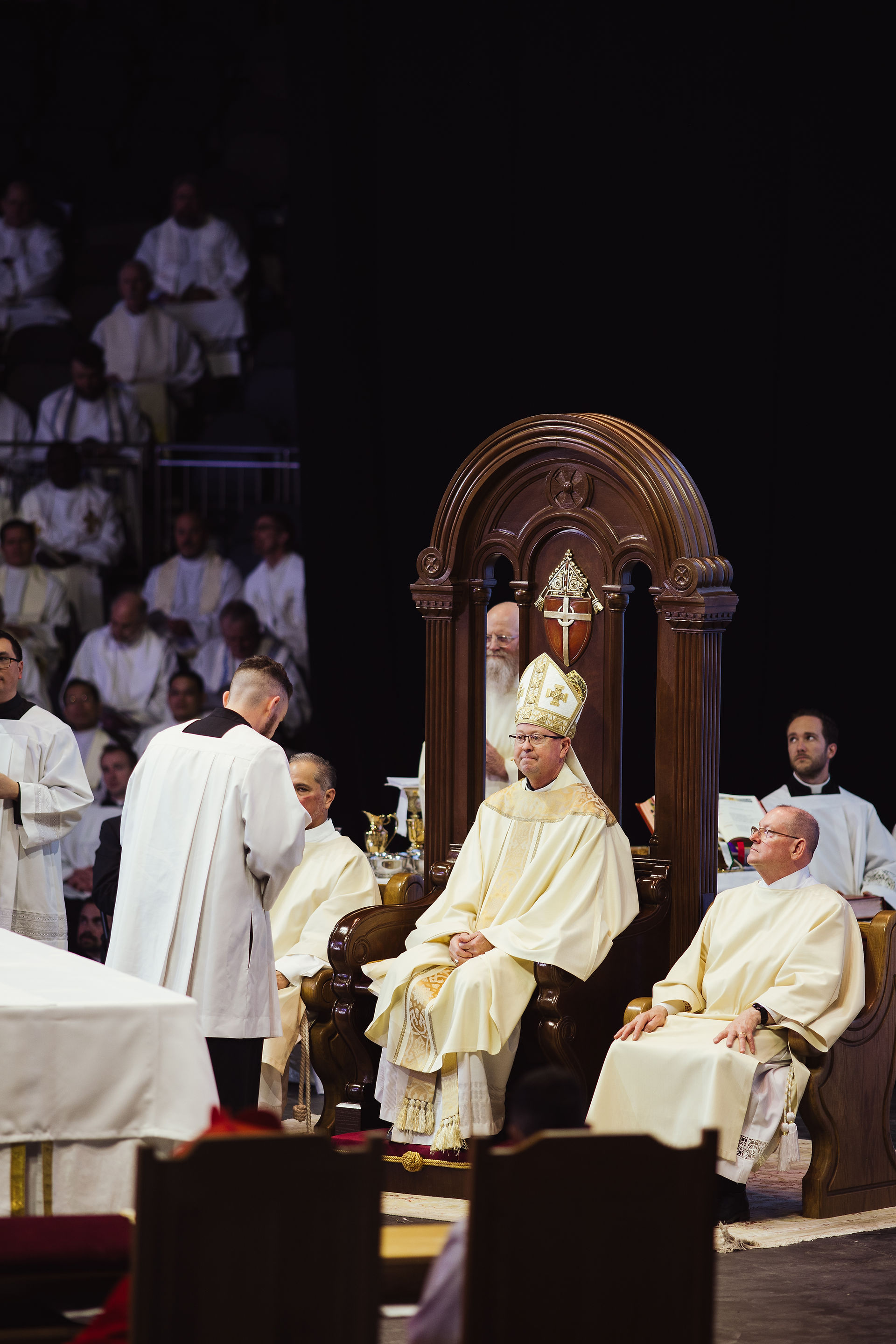 Clergymen in white robes at a ceremony. One sits on an ornate chair, wearing a mitre. Dark background, solemn mood, religious setting.