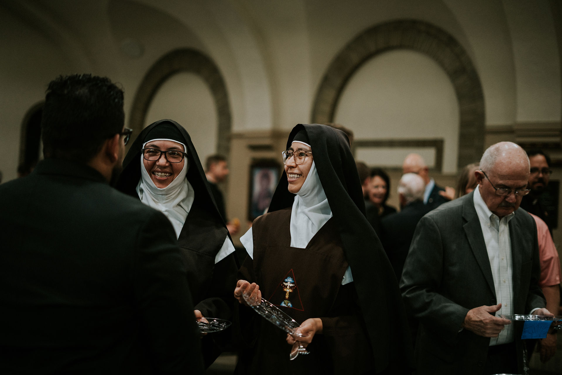Nuns in habits smile and hold trays at a social gathering in an arched hall, while a man in a suit looks at a paper. Warm, cheerful ambiance.