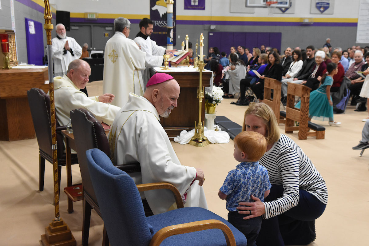 A bishop in white robes talks to a woman and boy in a church setting. A priest leads a service at an altar, with an audience seated behind.