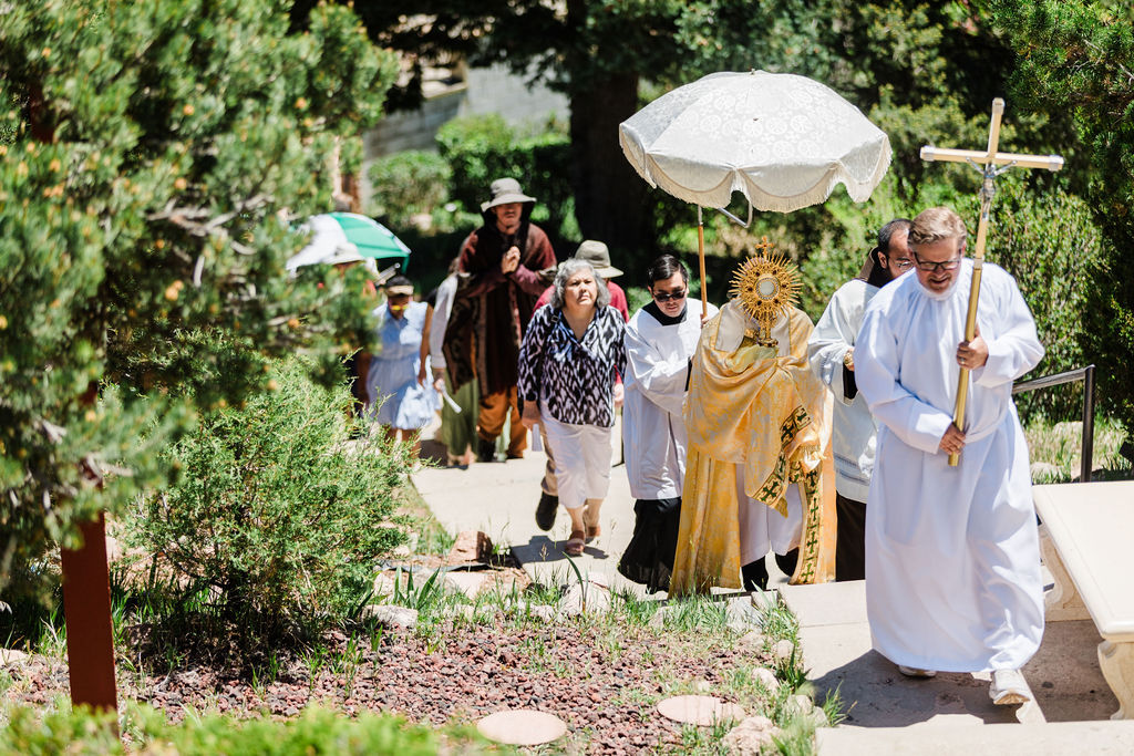 Outdoor Catholic procession climbs garden steps, priest carrying a cross beside a canopy over the Eucharist, with worshippers following.