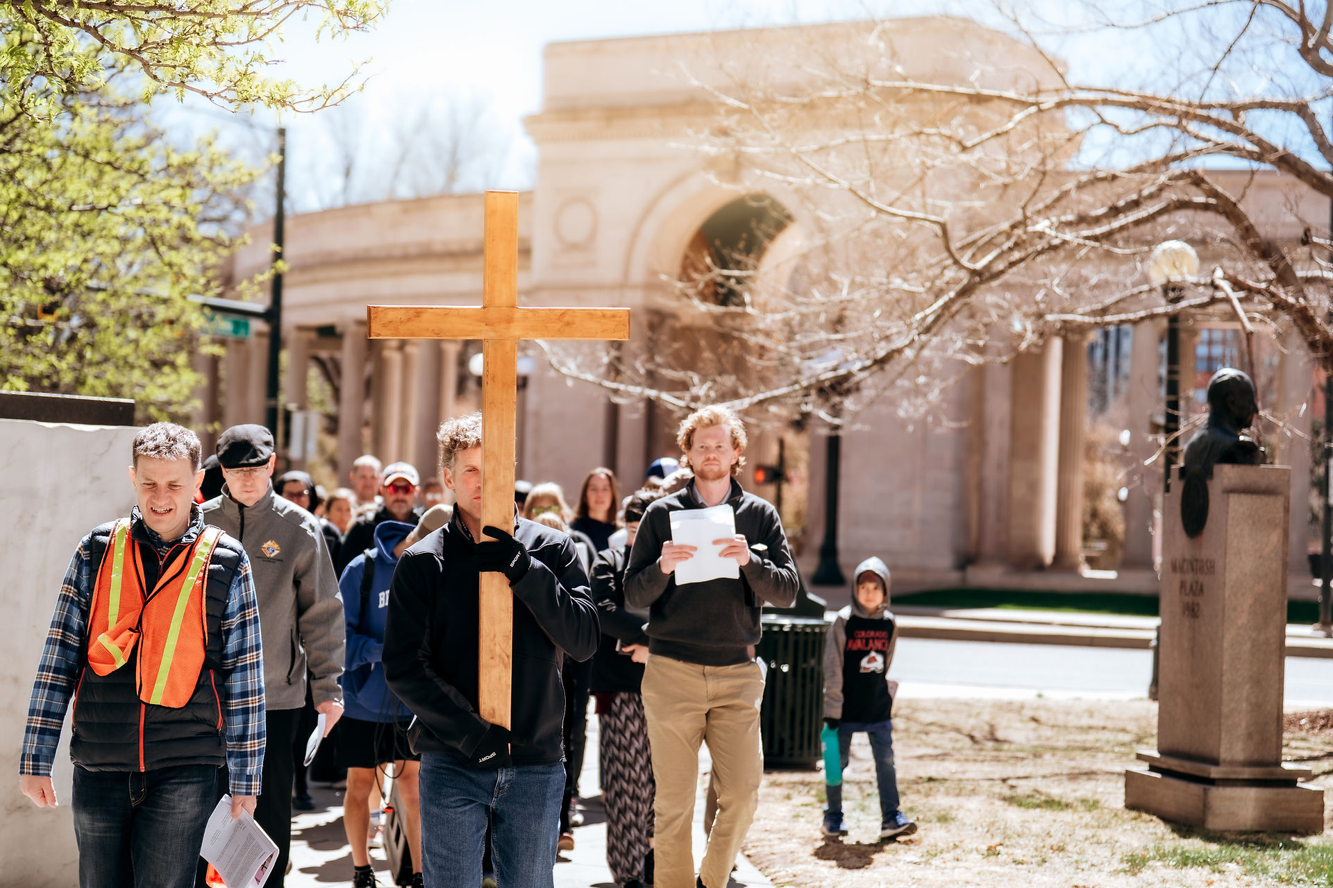 A man carries a wooden cross as participants follow behind, holding prayer sheets in a park near a monument.