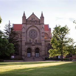 Stone church with red roof and rose window behind trees on a sunlit green lawn, calm and quiet scene