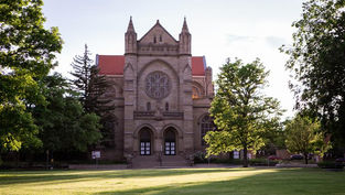 Stone church with red roof and rose window behind trees on a sunlit green lawn, calm and quiet scene