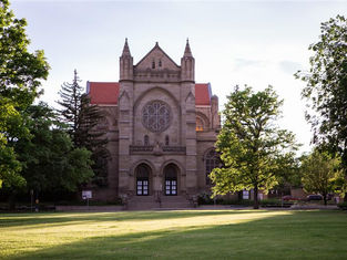Gothic-style church with red roof and large rose window. Surrounded by trees on a sunny day, casting soft shadows on the lush green lawn.