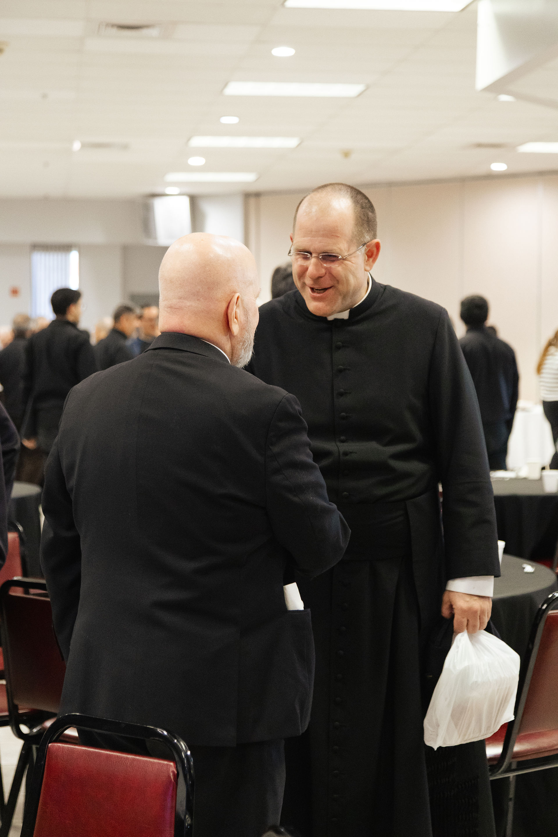 Two men in formal attire are having a friendly conversation in a conference room. One holds a white bag. The mood is warm and social.