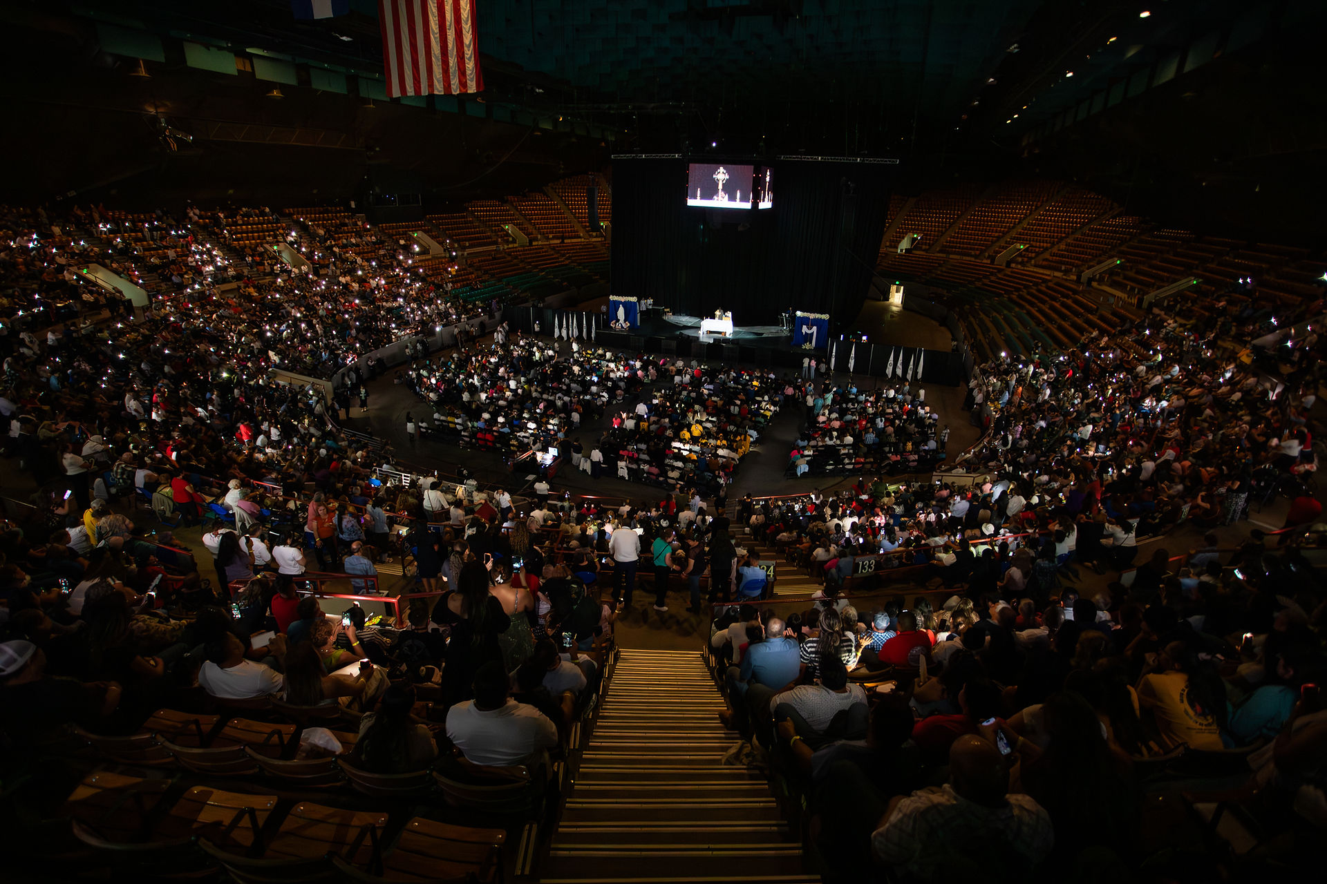 A packed indoor arena with a central stage and large screen showing a speaker; audience members are seated in rows, many holding up phones. An American flag hangs overhead.