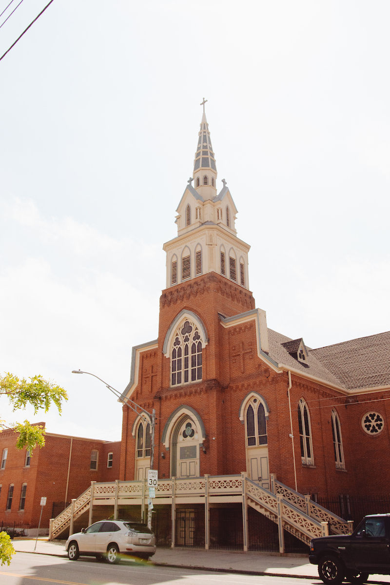 Red brick church with a tall steeple and cross, surrounded by a bright sky. Cars are parked nearby, with a visible "Speed 30" sign.