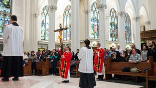 Clergy in red robes hold a large crucifix in a cathedral. Congregation stands in prayer. Stained glass windows in the background.
