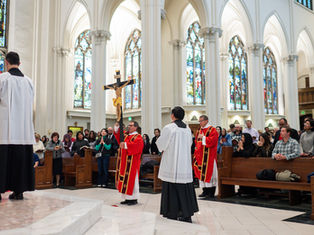 Clergy in red robes hold a large crucifix in a cathedral. Congregation stands in prayer. Stained glass windows in the background.