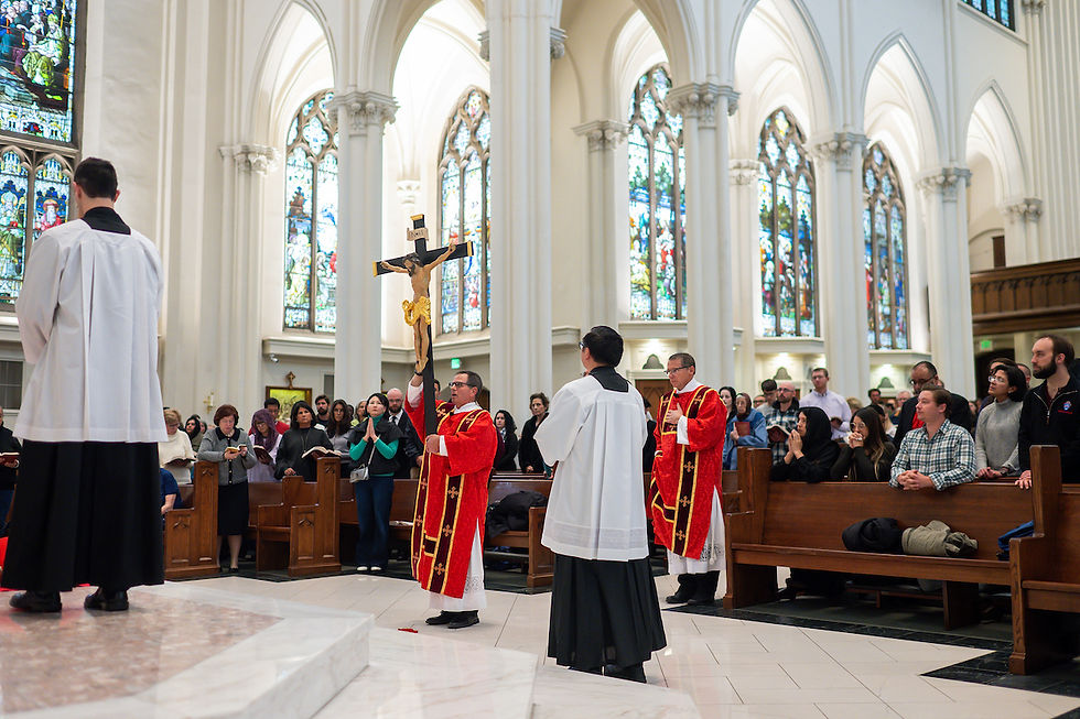 Clergy in red robes hold a large crucifix in a cathedral. Congregation stands in prayer. Stained glass windows in the background.