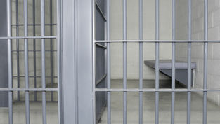 Empty jail cell with gray bars, a simple bed, and neutral walls, creating a stark and somber atmosphere. No text visible.