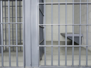 Empty jail cell with gray bars, a simple bed, and neutral walls, creating a stark and somber atmosphere. No text visible.