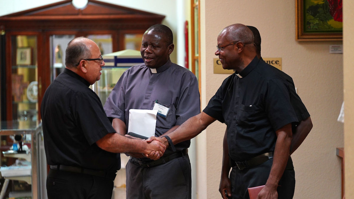 Three clergymen in black clerical attire greet and shake hands, smiling warmly. They're indoors, near a wooden cabinet and a sign reading "Entrance."
