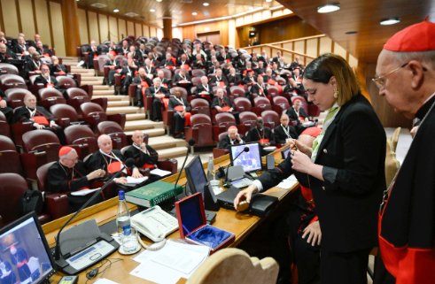 A woman stands near computers in a large, wood-paneled hall filled with cardinals in red and black attire. The mood is formal and focused.