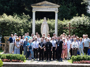 Group of tourists and priests pose before a white statue and cross in a sunlit garden.