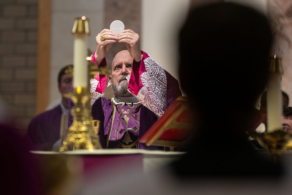 Bishop in purple vestments raises host during Mass. Candles and altar visible, solemn mood.