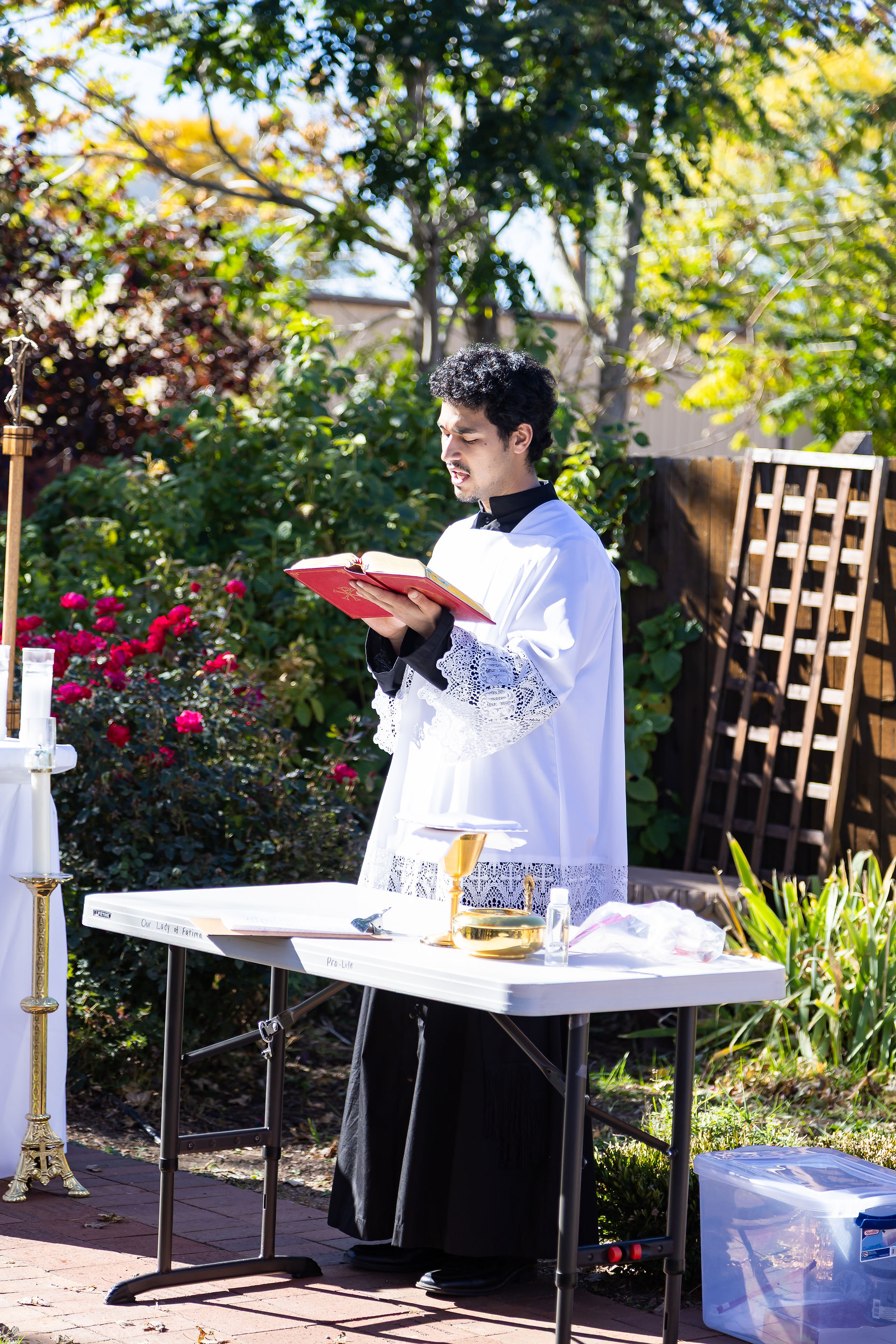 A man in white robes reads from a red book outdoors beside a table with chalice and candles. Greenery and flowers in the background.