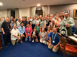 Large smiling group of adults, many with conference badges and clergy attire, posing together in a carpeted room with curtains and desk