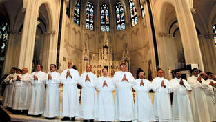 Deacons in white robes stand in a church, hands in prayer. Gothic arches and stained glass windows form the backdrop, creating a solemn mood.