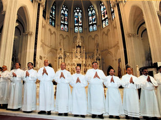 Deacons in white robes stand in a church, hands in prayer. Gothic arches and stained glass windows form the backdrop, creating a solemn mood.