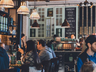 Cafe bustling with people chatting and using phones. Barista working at coffee machine under warm pendant lights. Espresso menu in background.