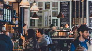 Cafe bustling with people chatting and using phones. Barista working at coffee machine under warm pendant lights. Espresso menu in background.