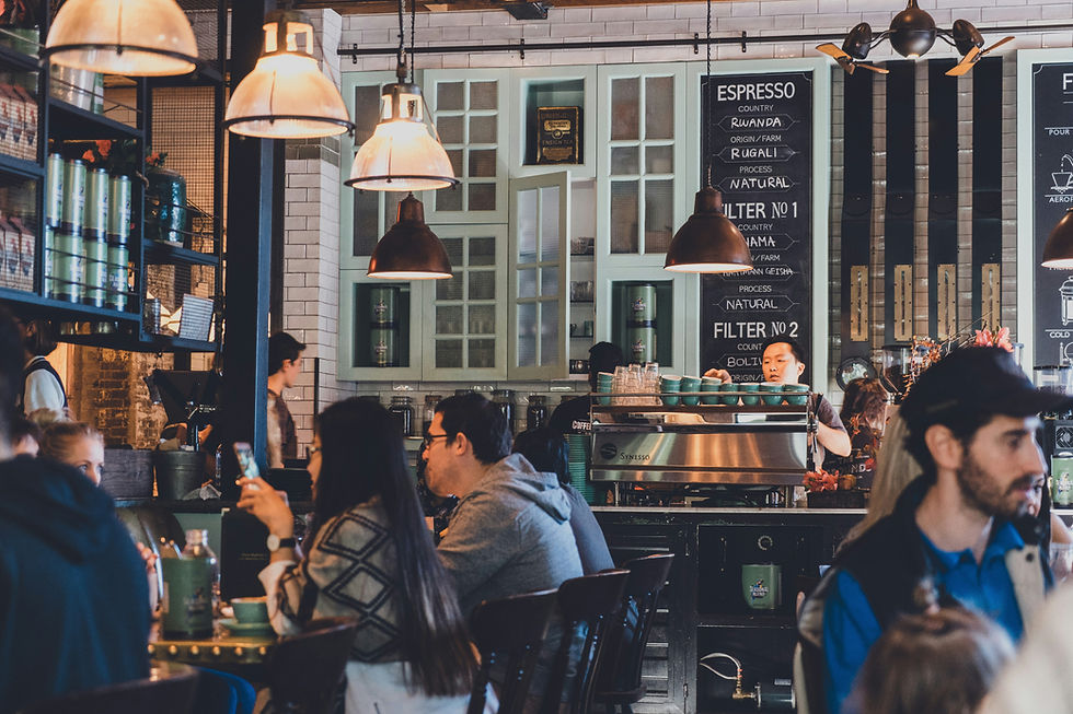 Cafe bustling with people chatting and using phones. Barista working at coffee machine under warm pendant lights. Espresso menu in background.
