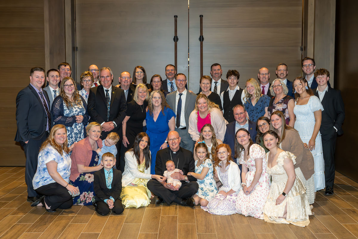 A large group poses indoors, some seated and some standing, on a wooden floor. People wear formal and floral outfits, smiling warmly.
