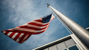 US flag waving on a tall flagpole against a blue sky, next to a modern building. The scene exudes a sense of patriotism and calm.