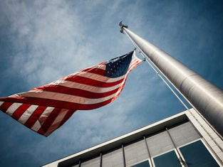 US flag waving on a tall flagpole against a blue sky, next to a modern building. The scene exudes a sense of patriotism and calm.