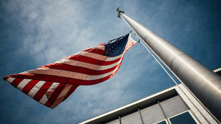 US flag waving on a tall flagpole against a blue sky, next to a modern building. The scene exudes a sense of patriotism and calm.