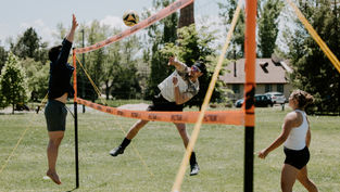 Three people play volleyball outdoors. One jumps to hit the ball over a net, another prepares to block. Trees and a house in the background.