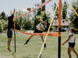 Three people play volleyball outdoors. One jumps to hit the ball over a net, another prepares to block. Trees and a house in the background.
