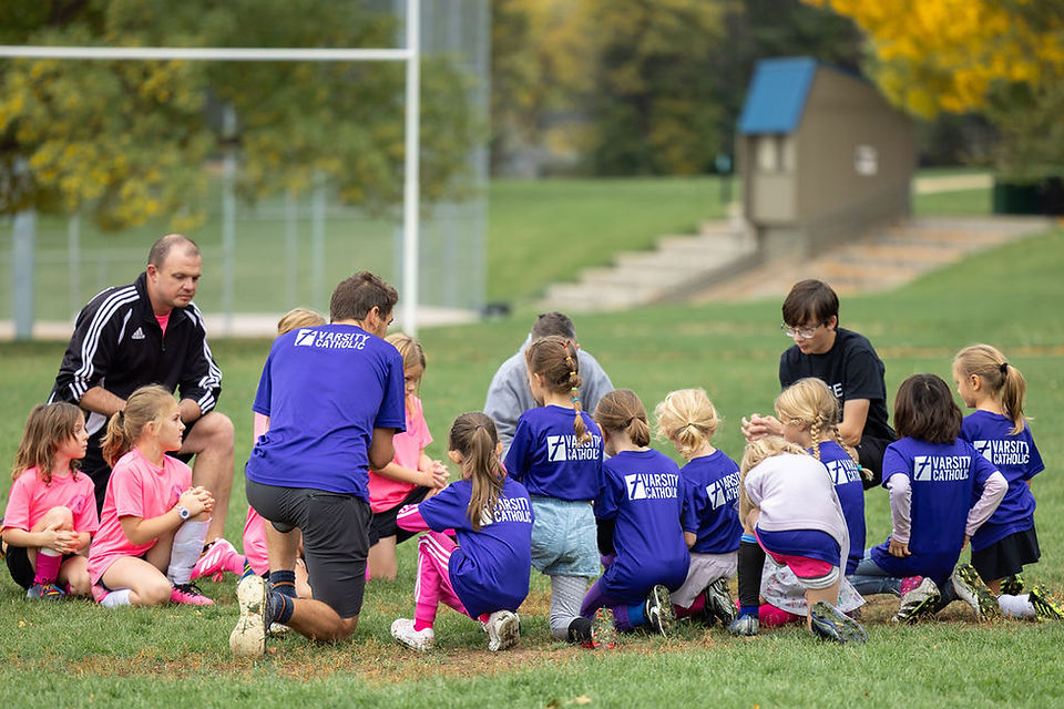 Youth soccer coaches and kids kneel in a huddle on a grassy field, wearing Varsity Catholic shirts, outdoors on a cool day