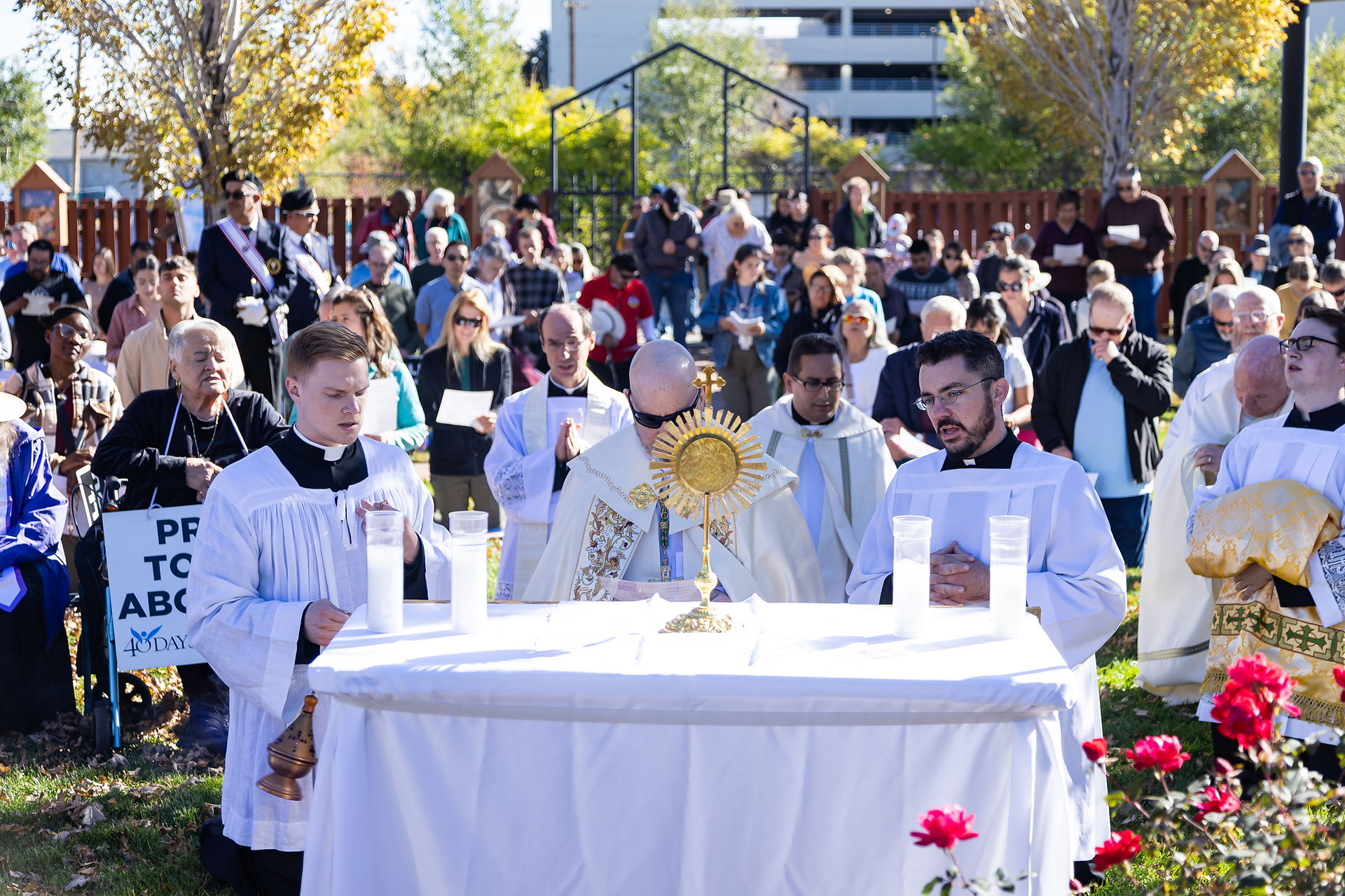 Archbishop and seminarians in white robes kneel at an altar with a golden monstrance outdoors. A crowd stands in prayer. Bright flowers and trees surround them.