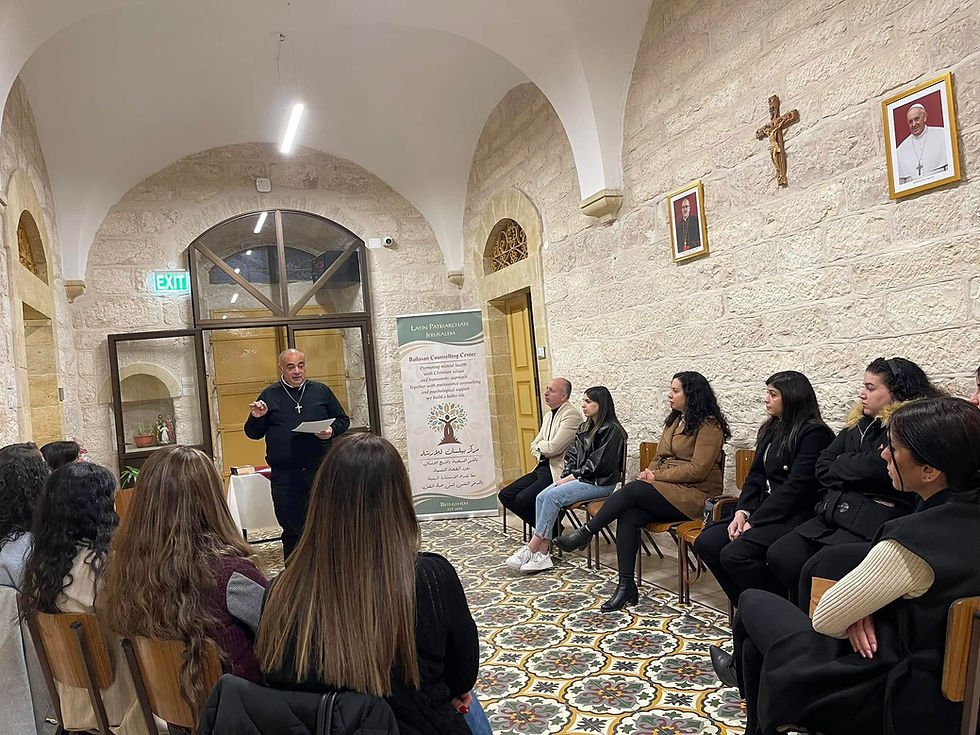 Priest lectures to seated group in stone room with archways. Colorful patterned floor, religious decor, and banner in background. Engaged atmosphere.