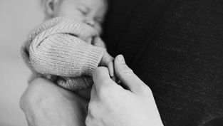Black and white image of a baby sleeping in a knitted sweater, holding an adult's hand, evoking a tender and peaceful mood.