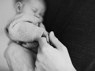 Black and white image of a baby sleeping in a knitted sweater, holding an adult's hand, evoking a tender and peaceful mood.