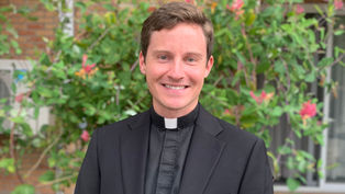 Smiling man in black clerical attire stands outdoors, vibrant green foliage with pink flowers in the background, conveying a cheerful mood.