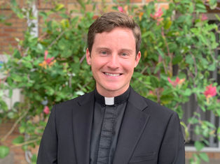Smiling man in black clerical attire stands outdoors, vibrant green foliage with pink flowers in the background, conveying a cheerful mood.